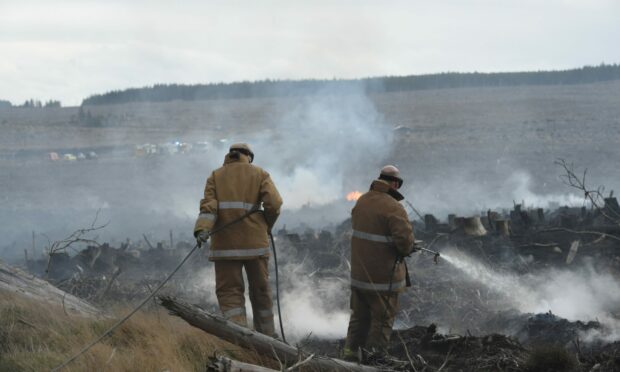 Two firefighters viewed from behind hosing down an area already damaged by a wildfire as smoke rises.
