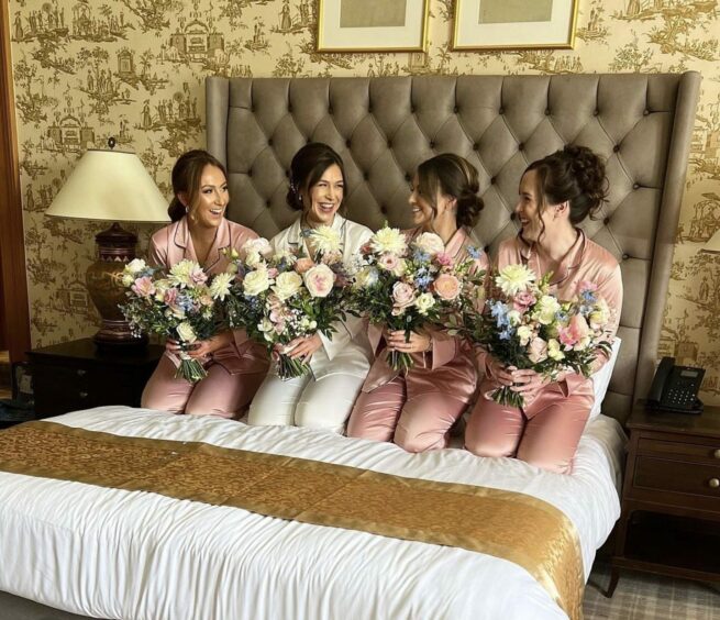 Three bridesmaids and a bride posing sat on a bed with their matching bouquets of flowers.