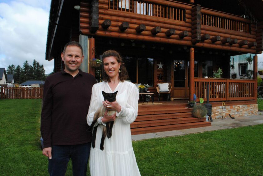 A man and woman with a cat standing in front of a log cabin