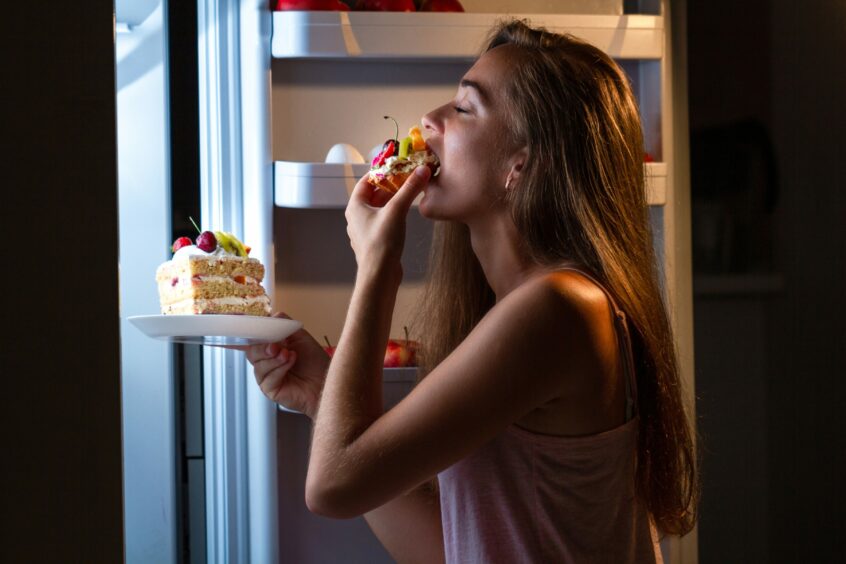 Hungry woman in pyjamas eating sweet cakes at night near refrigerator.