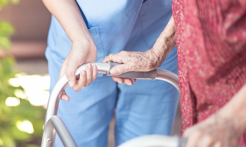 Care worker helps elderly woman on zimmer frame.