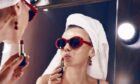 Woman wearing towel on head applying lipstick in a mirror.