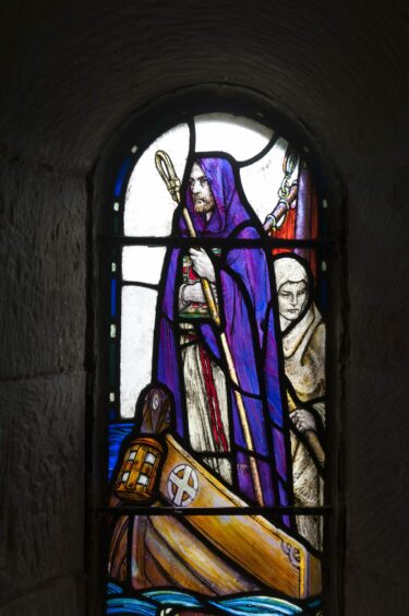 A stained glass window shoing St Columba in a purple cloak with a staff, standing in a boat