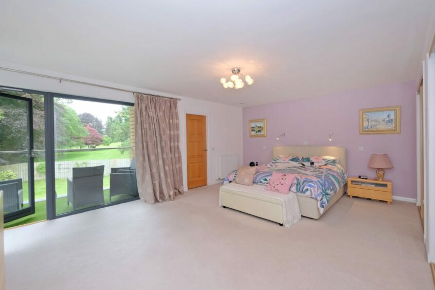 A bedroom with a double bed. This room has a pastel pink, purple and white colour scheme and a large window and patio doors that lead out to a balcony with chairs.