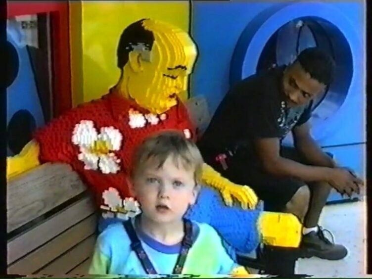 Still from a childhood clip of young Glen Cairns dressed in blue stands in front of a Lego man at Disney World Florida.