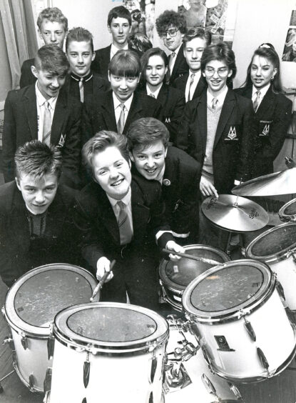 A group of students behind a drum kit, a girl sits at the drum kit with sticks