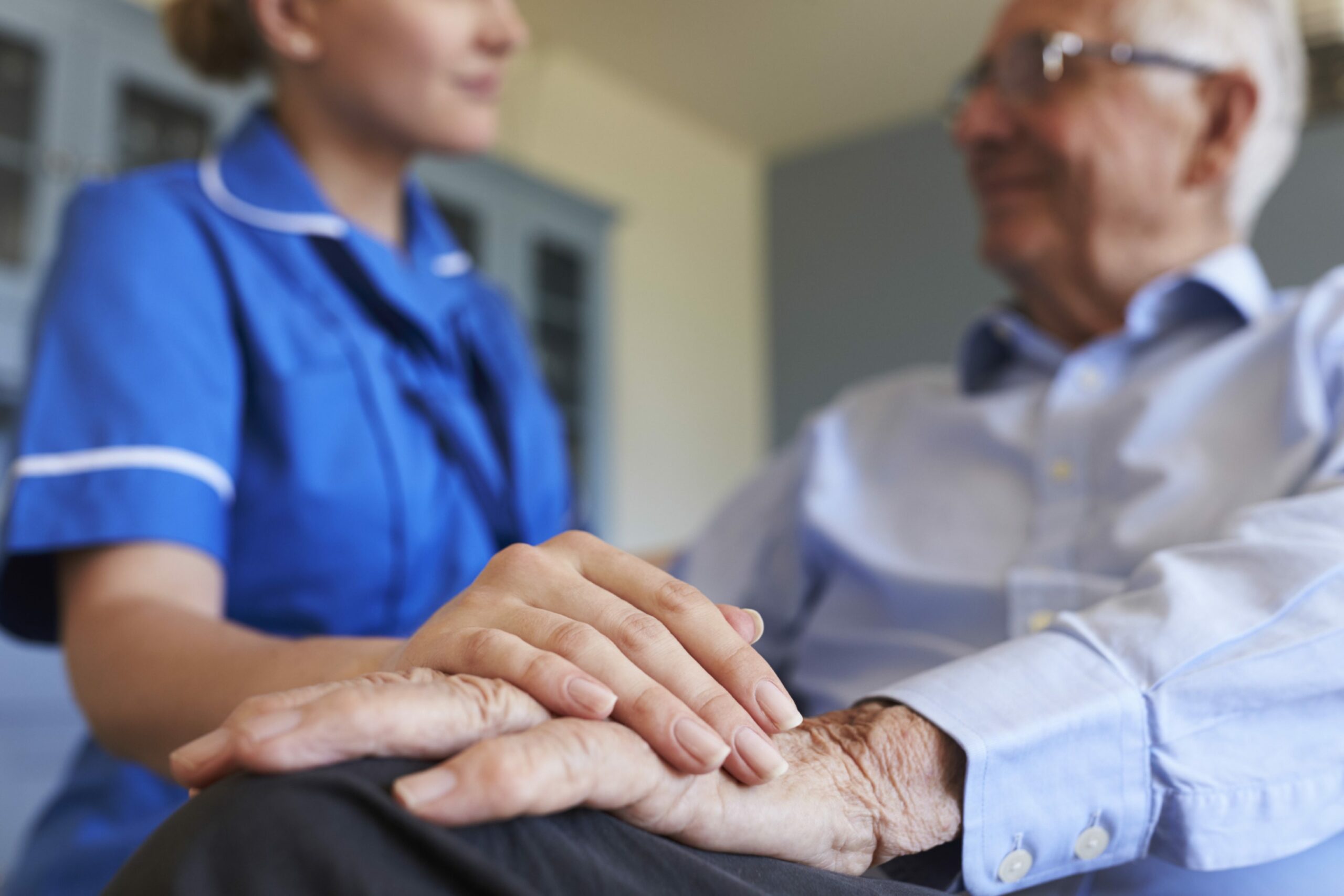 Carer standing up holding hand of old man sitting down. 