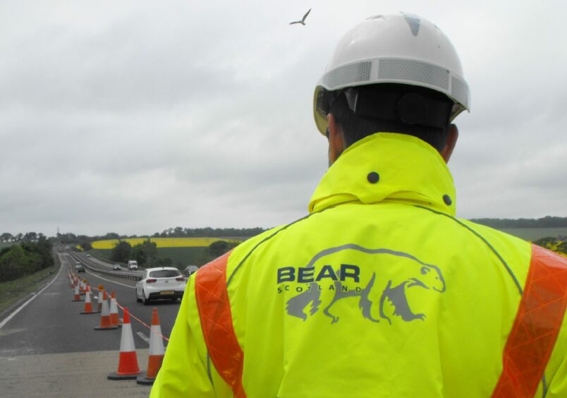 A Bear Scotland employee dressed in a high viability jacket and a hard hat standing on the side of the road. 