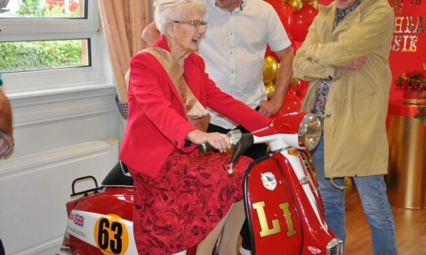 Jessie Clark laughing in a red dress while sitting on a bright red scooter.