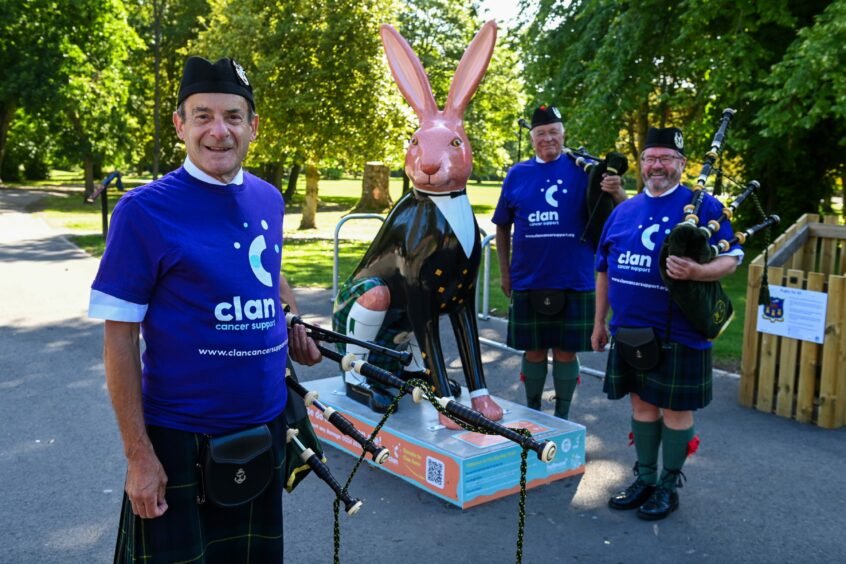 Gordon Caird (front), Tom Sinclair and Graeme Thom all stand beside a pink hare sculpture in Hazlehead Park, holding their bagpipes and wearing Clan's purple fundraising t-shirts.