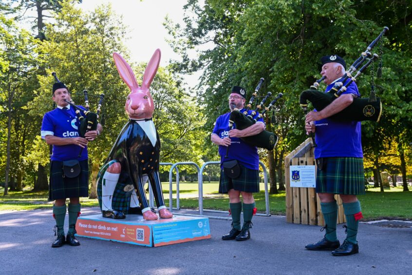 Gordon Caird (left), Tom Sinclair and Graeme Thom all stand beside a pink hare sculpture in Hazlehead Park, holding their bagpipes and wearing Clan's purple fundraising t-shirts.