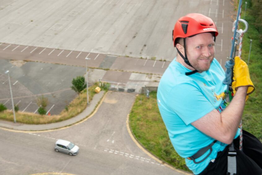 A man looks at the camera as he abseils with an empty car park in the background 