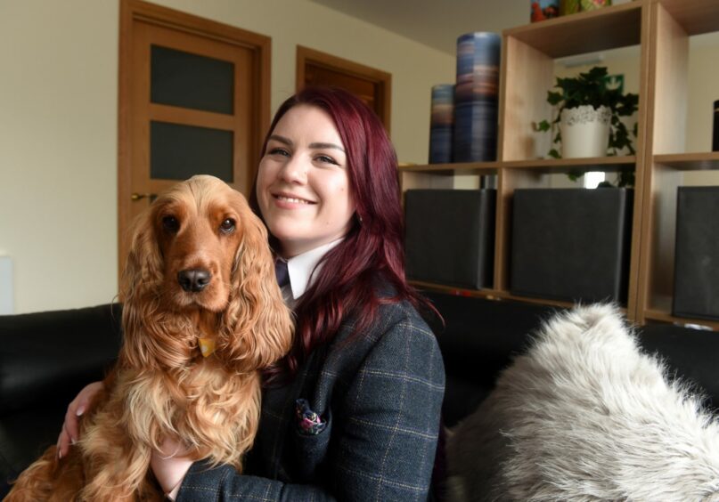 Embalmer Laura Wood and her dog Daisy. Laura is sitting on a sofa beside a fluffy cushion holding her dog. Laura wears tweed tartan and has long burgundy hair.