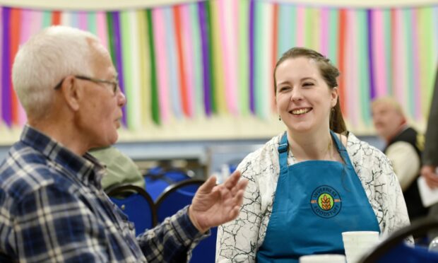 MSP Emma Roddick, dressed in a white blouse and blue apron, laughs with an Inverness resident at a table at Ness Bank Church.