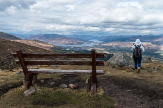Woman with backpack stood next to a bench over looking the Nevis Range.