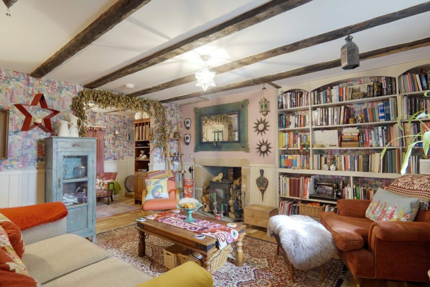 A living area in the Bleachfield house and cottage. The room features a wood burning fireplace, with a large built-in bookcase along the wall next to it. There is a mismatched sofa and armchairs with colourful cushions and throws, along with a wooden coffee table, a footstool, blue standing cabinet and a unique mirror on the wall. The walls are colourful and have plenty of decorations hanging up. There is an archway leading to another room and wooden beams along the ceiling.