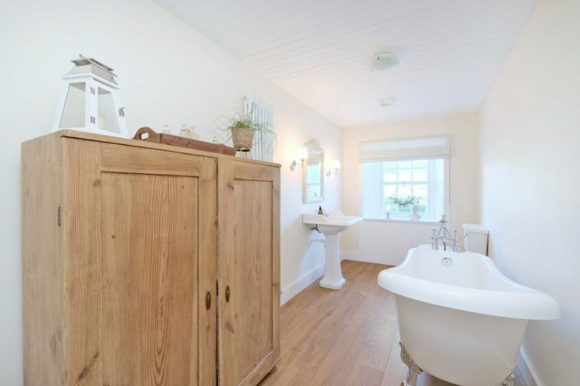 The bathroom in the cottage with light wooden flooring, a free standing cupboard and free standing bath. The toilet and sink are at the far side of the room next to the window