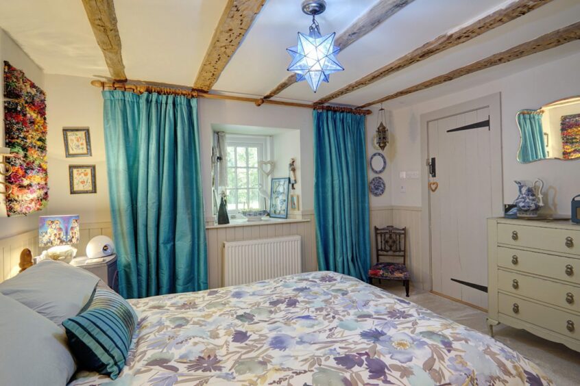A bedroom in Bleachfield House and Cottage with white walls and wooden beams along the ceiling. There's a window at the far side of the room with blue curtains, a large windowsill and a radiator underneath. There's a double bed with a blue floral bedspread, bedside table and chest of drawers. On the wall above the bedside table is a panel of colourful faux flowers
