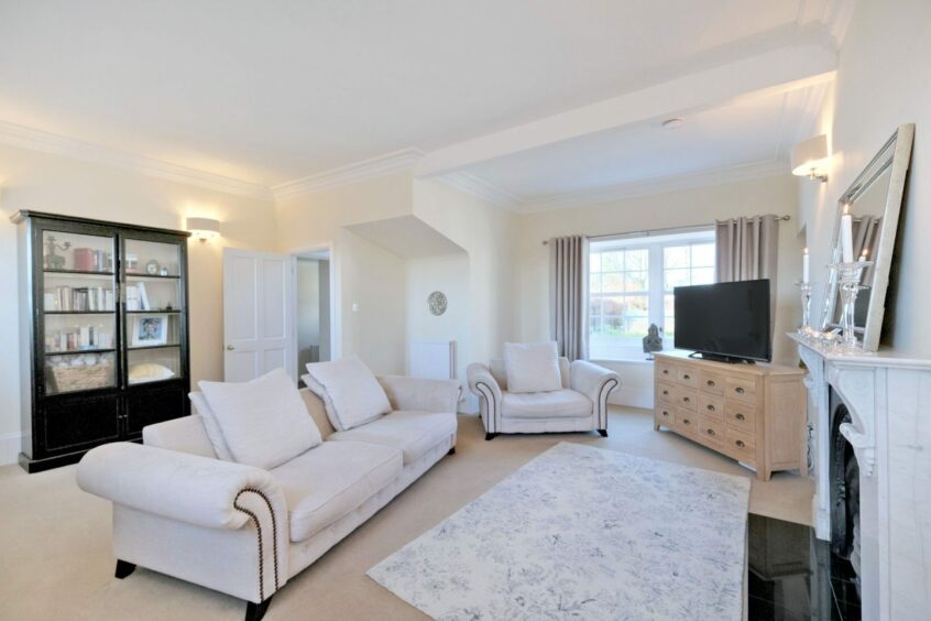 The living area of the property, with a white sofa and armchair, a black and white fireplace with a mirror on the mantelpiece, a black bookcase and a pale wooden unit with the flat screen television on top