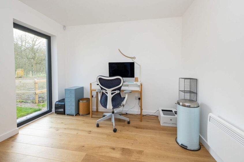 The home office has a white desk with wooden legs, a modern desk chair and file storage drawers and shelves. There's a large window to the left of the desk providing plenty of natural light