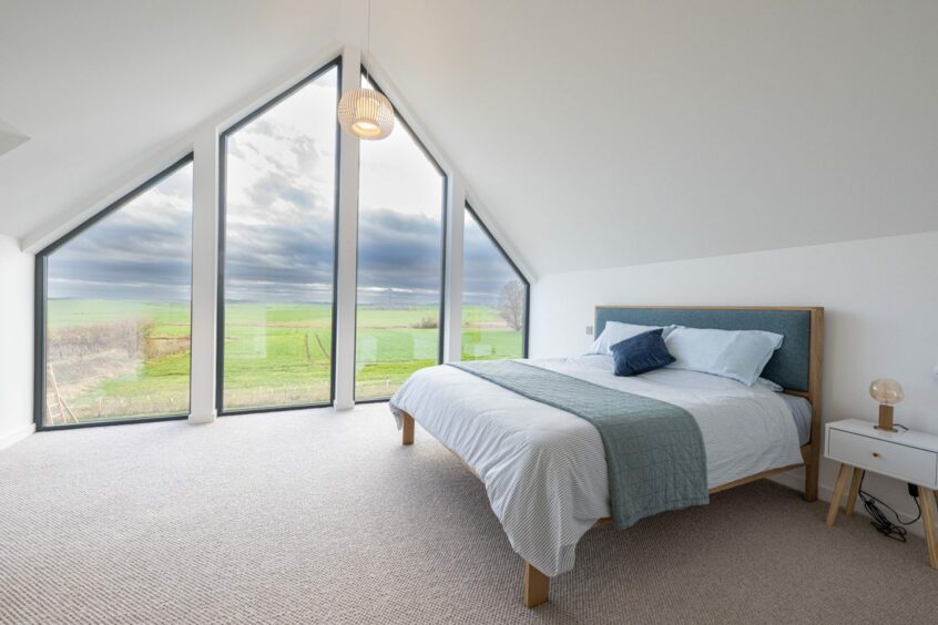 One of the bedrooms in the Udny house, with four large windows reaching to the roof of the house and overlooking the Aberdeenshire countryside. The walls and bedsheets are white, the headboard, throw and cushions on the bed are shades of blue. There's a small bedside table with a lamp on it.