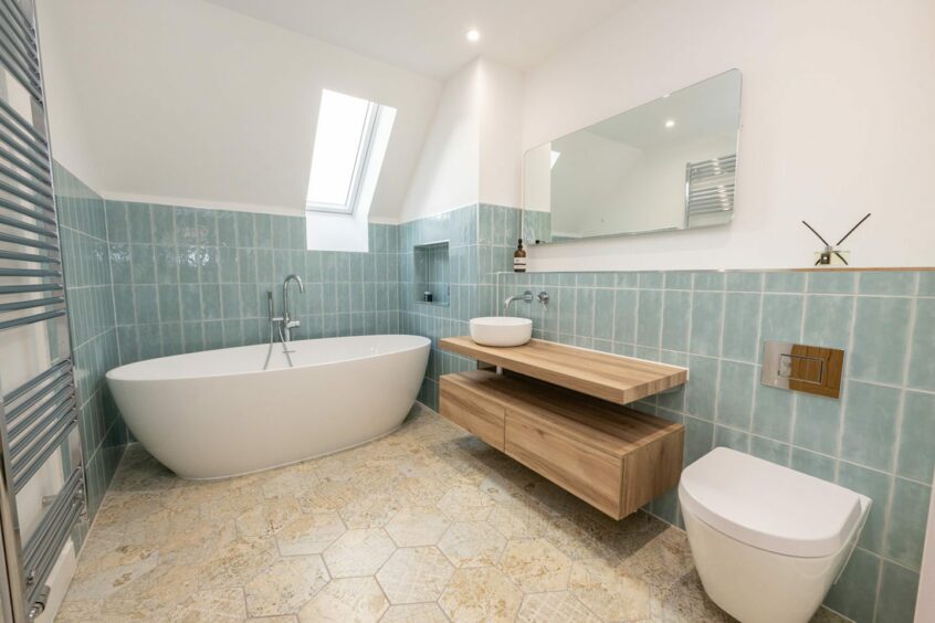 The bathroom has plenty of room. The upper half of the walls are painted white with the lower half covered in pale blue rectangular tiles. The sink is a white basin on a wooden base with two drawers for toiletries. There's a modern-looking bath at the far side of the room with an alcove in the wall perfect for scented candles and toiletries. There's a shelf running along the wall above the sink and toilet with soap and incense on it. The flooring is made up of textured beige octagonal tiles.