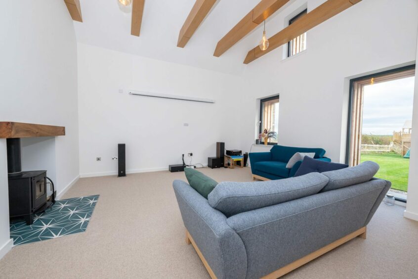 A living area with a woodburner fireplace, two blue sofas and a projector screen and sound system at the far side of the room. The walls are a crisp white, with three large windows with views over the Aberdeenshire countryside.