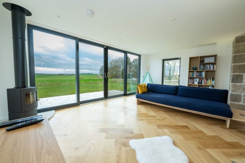 The main living area in the Udny home, in the same open-plan room as the kitchen and dining areas. The room features laminate wooden flooring, a modern dark blue sofa with a wooden base, a large bookcase and woodburner. There's a large set of glass patio doors looking out at the Aberdeenshire counrtyside, that lead out to the garden.
