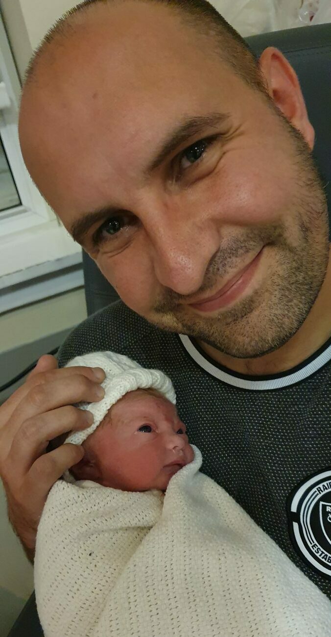 David Mackay smiling at the camera while holding newborn baby wrapped in white blanket and wearing hat. 