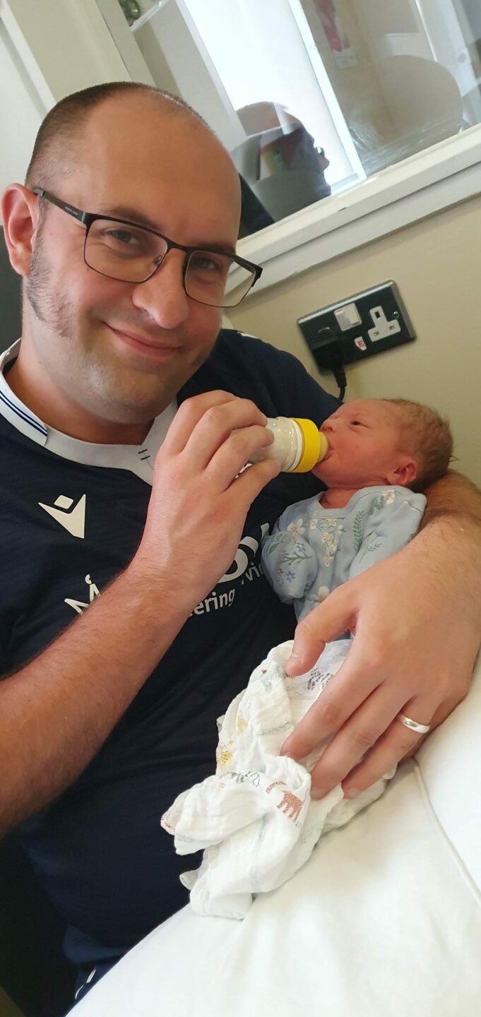 David Mackay feeding daughter Isla with a bottle while looking at camera. 