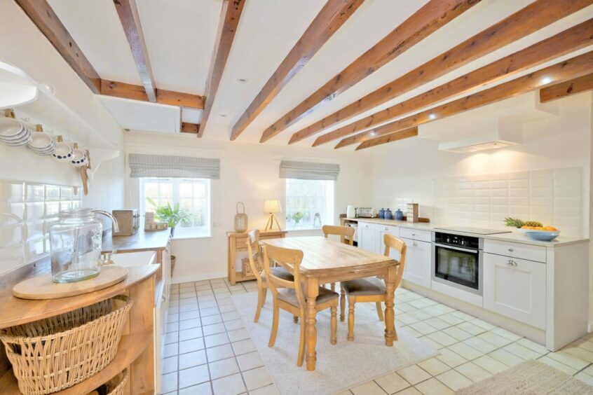 Eden Cottage near inverurie has a spacious kitchen with a dining table seating four in the middle of the room. The kitchen is primarily white with light wooden accents, including wooden beams along the ceiling.