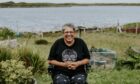 A woman using an electric wheelchair smiles in a garden with a loch in the background.