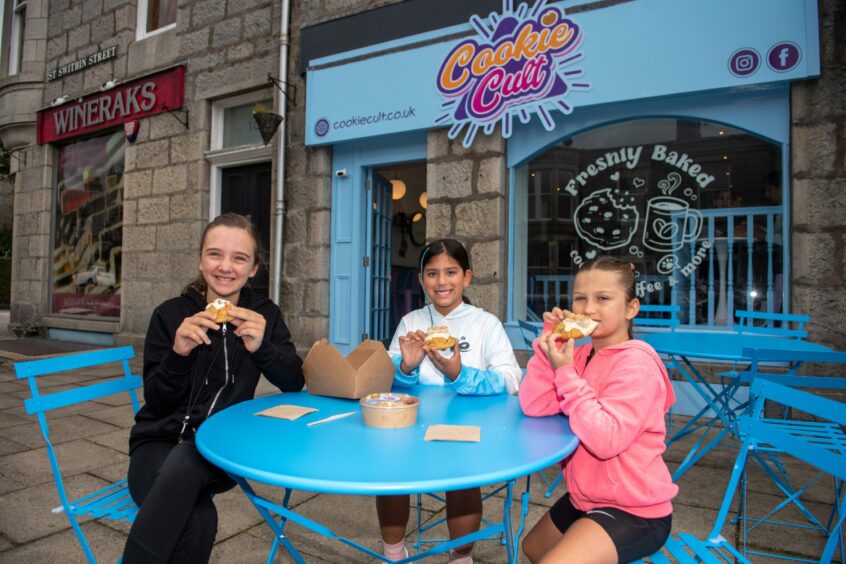 Three girls sitting outside the shop, tucking into their cookies
