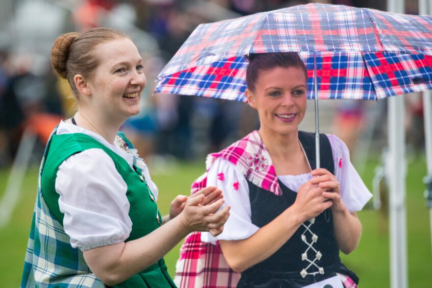 Two Highland dancers smile as they look ahead. One wears a green waistcoat and kilt while the other girl, under a red and blue umbrella, wears a pink kilt.