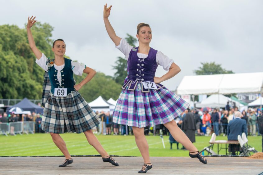 Two Highland dancers on stage. The front dancer wears a purple kilt and waistcoat, while the second at the back is in green.