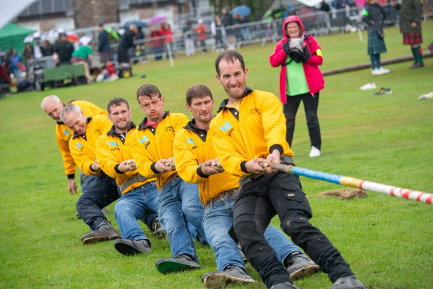 Six members of the Cornhill tug o'war team pull on the rope. They wear the team's mustard yellow rugby shirt, jeans and boots as they dig into the ground, pulling the rope.