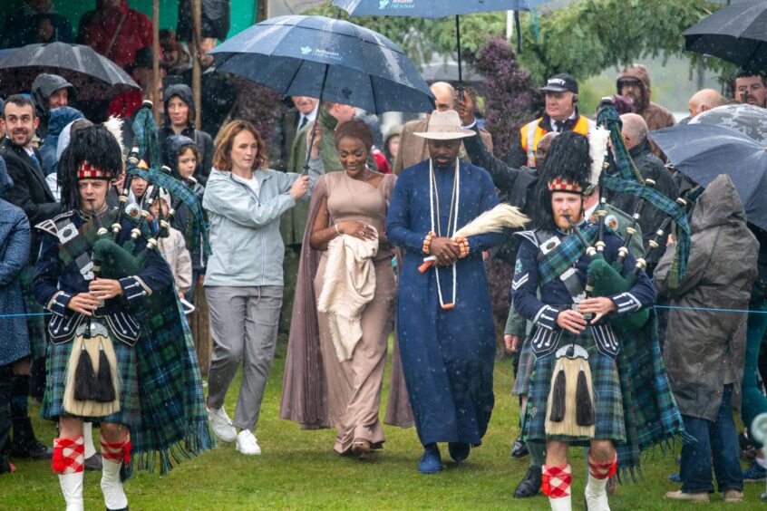 Two pipers, in formal Scottish attire, lead the King and Queen of Warri onto the field. The queen wears a long blush coloured dress while the king has a navy robe and hat on. They are walking under umbrellas through the crowd.