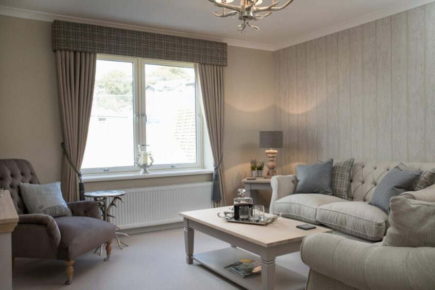The living room of one of the Inverurie bungalows - with light cream walls and couches. There's a brown arm chair with a small end table and a white coffee table in the centre of the room
