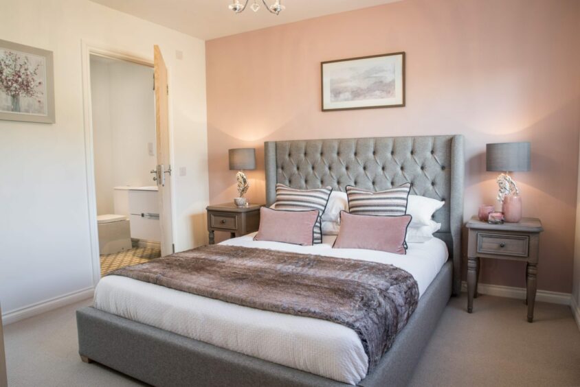 One of the bedrooms, with white walls, a pink feature wall, a plush grey bed frame and headboard, a cream carpet and two bedside tables with lamps.