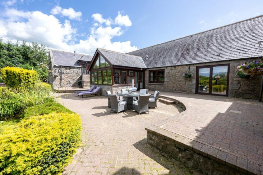 The bricked patio area, with a large section of greenery next to it. There are two sun-loungers, a table and six chairs to enjoy the outdoors