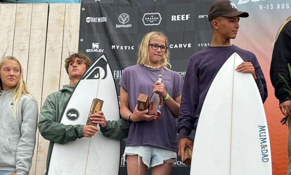 Teenagers (two girls/two boys) stand on a podium. Both the boys are holding surfboards.