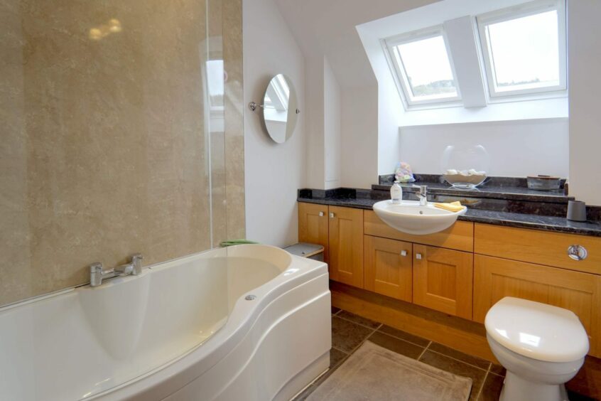 The bathroom with cream walls, a curved bath with glass screen, wooden cabinets with black countertops and two small windows on the slanted section of the ceiling
