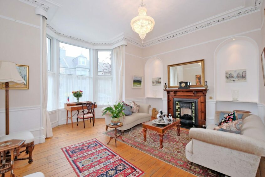 A living area in the west end property in Aberdeen. The room has white and cream walls and an intricate trim around the ceiling. Two cream sofas are on either side of the fireplace, with a coffee table in the middle. A large bay window provides the room with plenty of light, with a wooden writing desk and chair in front of it.