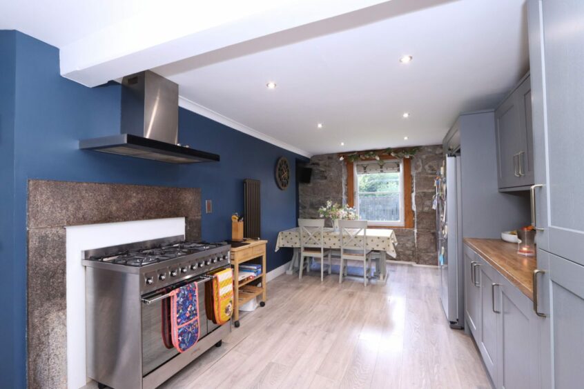 The kitchen in the royal deeside cottage with grey-blue cupboards, wood-effect countertops, a metal oven with a gas hob and a dining table for four. The walls are painted blue except one wall with exposed stone, which features a window