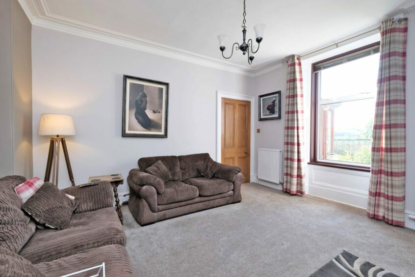 The living room has a plush grey carpet, two dark fabric couches, a large window and plaid curtains