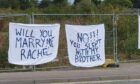 Two bedsheets attached to fencing side-by-side. One saying "Will you marry me Rachel?", one on right saying "No! You slepy with my brother."