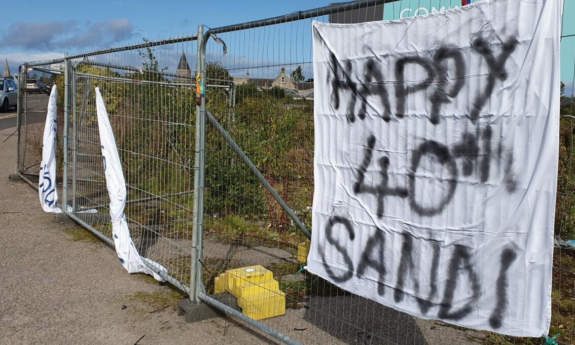 Image of fencing at Edgar Road roundabout with bedsheet saying "happy 40th sandi" and two others fallen to ground. 