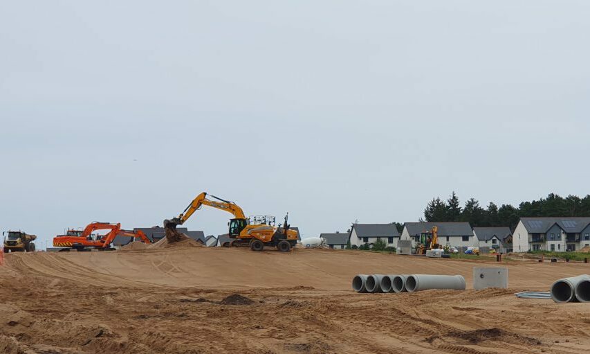 Construction vehicles on large sandy land with homes in background. 