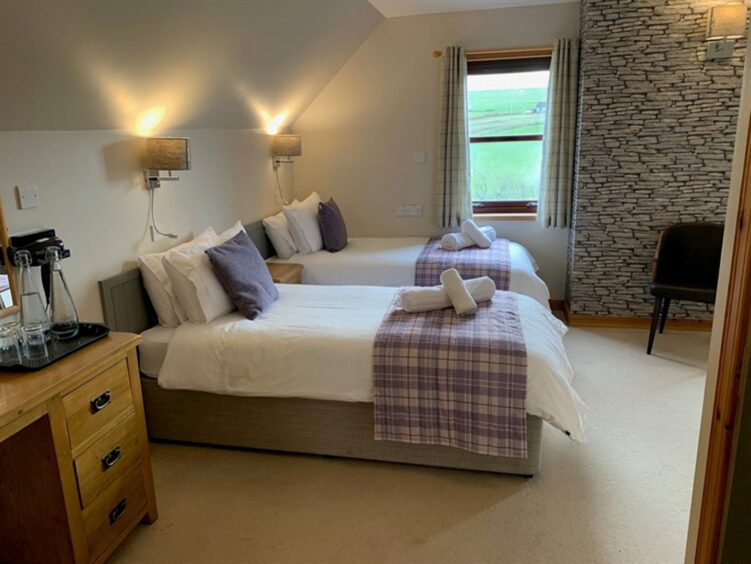 The Standing Stones Hotel bedroom, with two twin beds, a wooden bedside table between them, a wooden dresser, a cream carpet and two wall lamps above the beds. There's a window overlooking the Orkney countryside next to one of the beds
