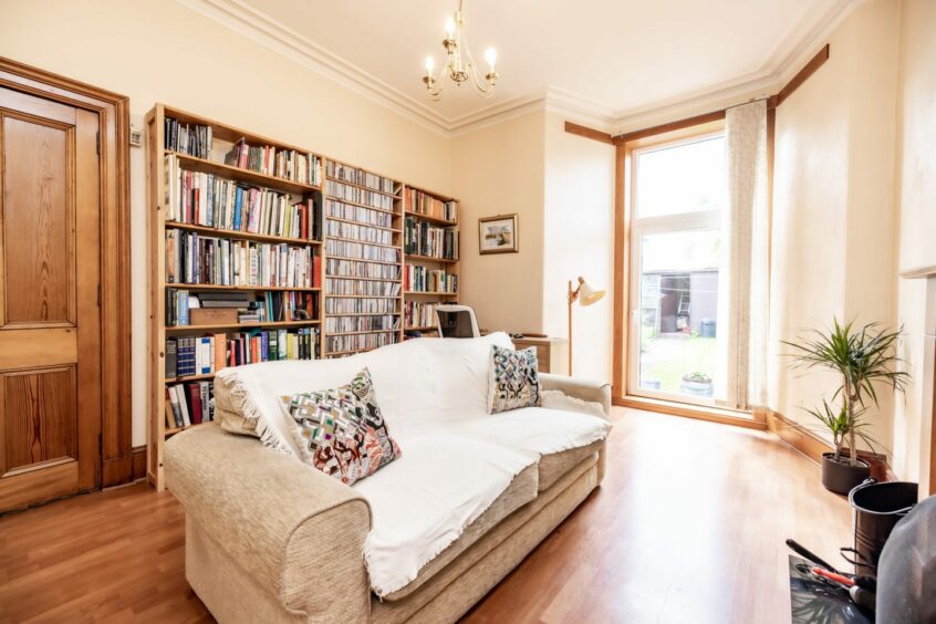A study in the period property in Aberdeen, with a large bookcase spanning from the door to the window, a desk and chair, a floor lamp and a plush cream sofa with cushions and a throw across from a fireplace